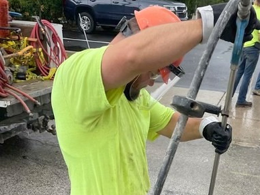 A Darr's crew member in a hard hat and high-visibility shirt working a locating rod at an opening in the asphalt, with the truck and its hose reels behind