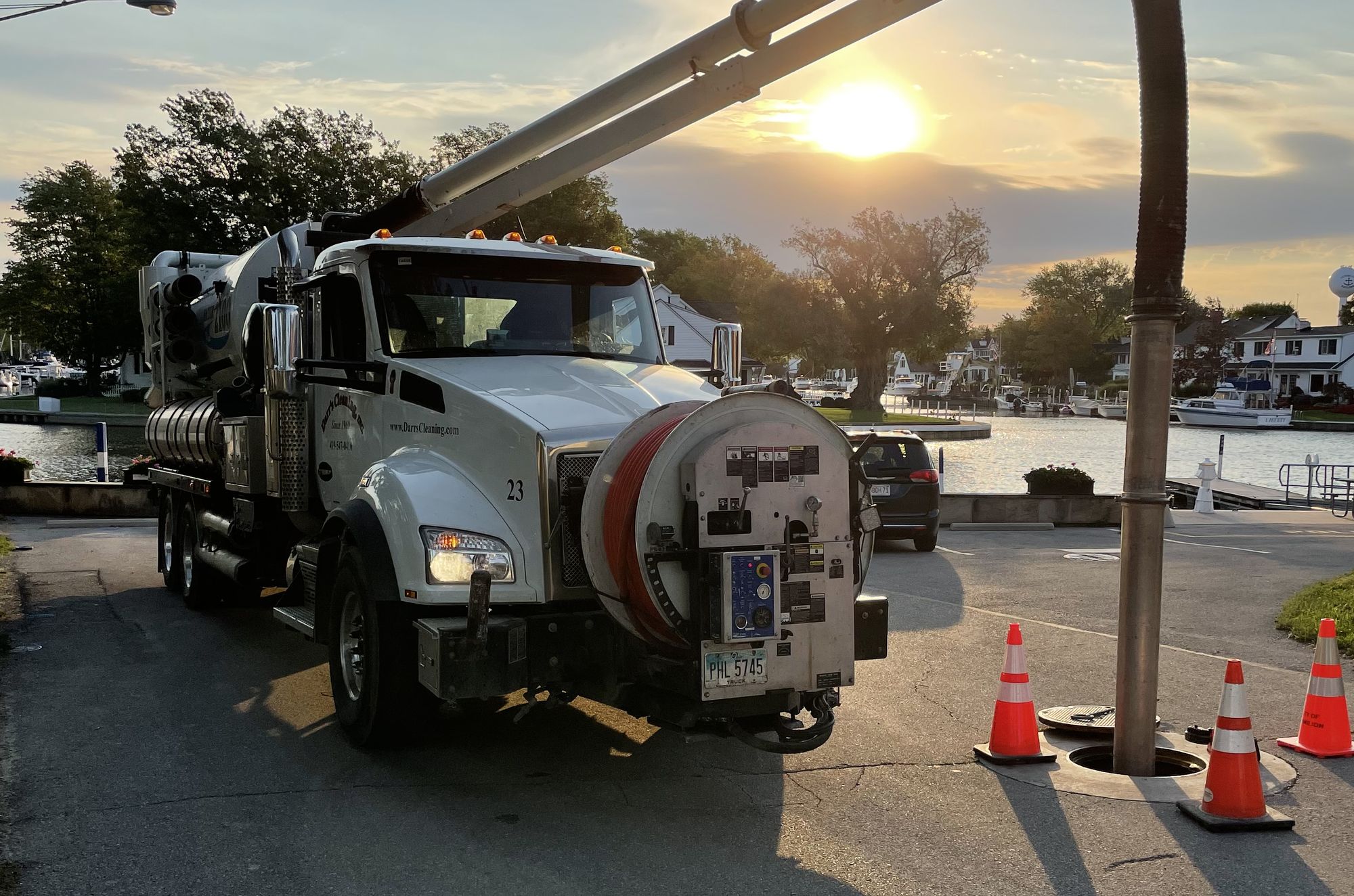 The vacuum tube of a Darr's Cleaning combination truck running down into an open sewer manhole at sunrise, traffic cones set around the opening, a marina behind